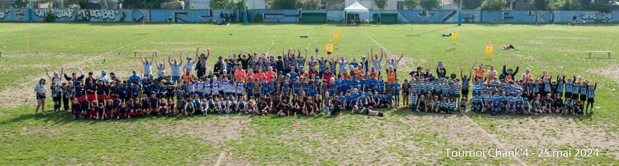 Une photo qui montre plusieurs équipes de jeunes de moins de 10 ans et leurs entraîneurs sur un terrain herbeux lors de l'événement « Tournoi Chank'4 », le 25 mai 2024. Les joueurs sont vêtus d'une variété de maillots colorés, représentant différentes équipes. Ils sont disposés en rangées, certains assis et d’autres debout, tous levant les bras dans une pose de célébration. Le terrain présente des signes d'usure, avec des taches de saleté au milieu de l'herbe, soulignant son utilisation active. L'ambiance générale est festive et énergique, reflétant la joie et l'excitation du tournoi.